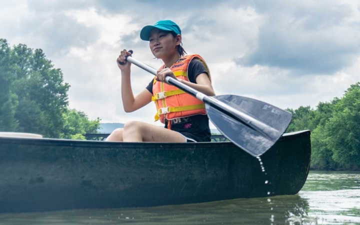 A young person wearing a life jacket paddles a canoe on calm water. There are trees in the background. 